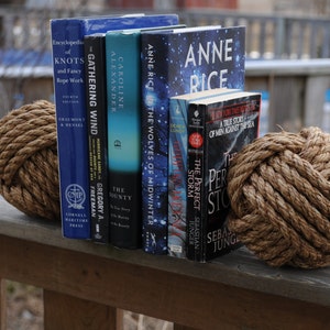 K&ouml;nnte beinhalten: Eine Anordnung von B&uuml;chern mit blauen, t&uuml;rkisfarbenen und schwarzen Einb&auml;nden, die von zwei dekorativen Seilknoten-Kugeln gehalten werden. Die B&uuml;cher tragen Titel wie "Encyclopedia of Knots" und "Anne Rice". Die Kugeln sind aus dickem, gedrehtem braunem Seil gefertigt.