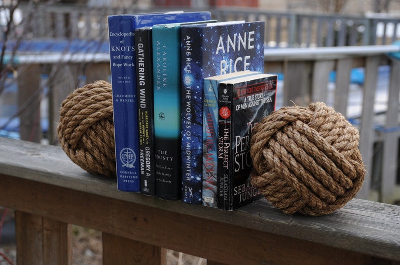 K&ouml;nnte beinhalten: Eine Anordnung von B&uuml;chern mit blauen, t&uuml;rkisfarbenen und schwarzen Einb&auml;nden, die von zwei dekorativen Seilknoten-Kugeln gehalten werden. Die B&uuml;cher tragen Titel wie "Encyclopedia of Knots" und "Anne Rice". Die Kugeln sind aus dickem, gedrehtem braunem Seil gefertigt.