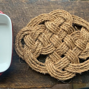 May include: A red ceramic baking dish with white interior and a natural fiber rope trivet. The trivet is woven in a knot pattern.