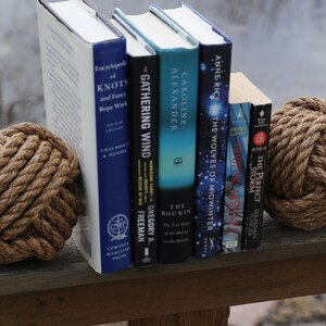 K&ouml;nnte beinhalten: Eine Pr&auml;sentation von B&uuml;chern und dekorativen Seilknoten. Die B&uuml;cher haben verschiedene Titel und Farben, darunter "Encyclopedia of Knots" und "The Gathering Wind". Die Seilknoten sind braun und f&uuml;gen ein maritimes Thema hinzu.