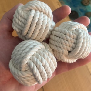 May include: Three white rope balls, knotted together, are held in a hand. The balls are made of thick, white rope and are about the size of a tennis ball.