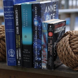 K&ouml;nnte beinhalten: Eine Sammlung von B&uuml;chern mit nautischen Themen, darunter Titel wie "Knoten und ausgefallene Seilarbeiten" und "Der perfekte Sturm", wird zwischen zwei dekorativen Seilkugeln pr&auml;sentiert. Die B&uuml;cher sind auf einer Holzoberfl&auml;che angeordnet.