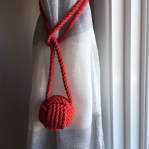 May include: Red rope curtain tieback with a knotted ball at the end. The tieback is wrapped around a white sheer curtain.