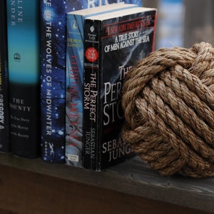 K&ouml;nnte beinhalten: Ein dekorativer Seemannsknoten aus dickem, braunem Seil liegt neben einer Reihe von B&uuml;chern. Die B&uuml;cher tragen Titel wie "The Perfect Storm" und "The Wolves of Midwinter". Die Szene erinnert an ein maritimes oder K&uuml;sten-Thema.