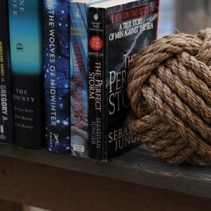 K&ouml;nnte beinhalten: Ein dekorativer brauner Seilknoten neben einer Reihe von B&uuml;chern. Zu den Titeln geh&ouml;ren "The Perfect Storm" und "The Wolves of Midwinter". Die B&uuml;cher stehen auf einer Holzoberfl&auml;che.