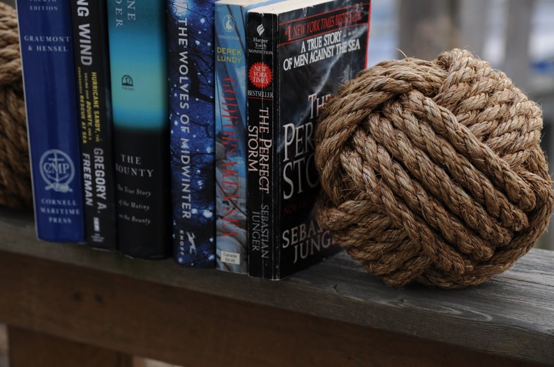 K&ouml;nnte beinhalten: Ein dekorativer brauner Seilknoten neben einer Reihe von B&uuml;chern. Zu den Titeln geh&ouml;ren "The Perfect Storm" und "The Wolves of Midwinter". Die B&uuml;cher stehen auf einer Holzoberfl&auml;che.