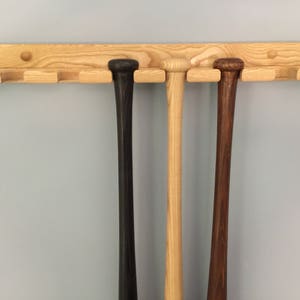 May include: A wooden baseball bat rack with six slots, mounted on a wall. Three bats are displayed in the rack, one black, one natural wood, and one brown.