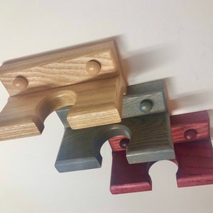 May include: Three wooden wall mounted gun racks in natural, gray, and red stained finishes. Each rack has a rounded cutout for holding a gun barrel and two small round knobs.