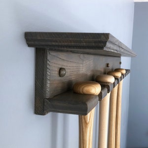 May include: A gray wooden baseball bat rack with a shelf on top. The rack has six slots for bats and is mounted on a wall. There are four bats in the rack, all with natural wood finishes.