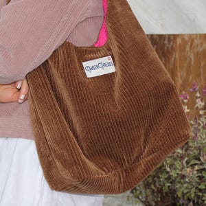 May include: A brown corduroy tote bag with a pink accent and a rectangular label. The bag is worn over the shoulder. The image also shows a pink corduroy jacket and a white skirt.