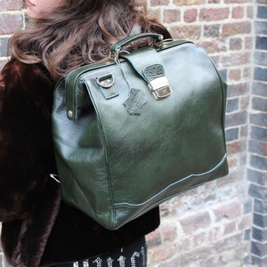 Puede incluir: Un maletín de médico de cuero verde oscuro con un asa superior y un cierre metálico. La bolsa tiene forma rectangular y se lleva como mochila. La bolsa está en la espalda de una persona que lleva un abrigo de piel marrón.