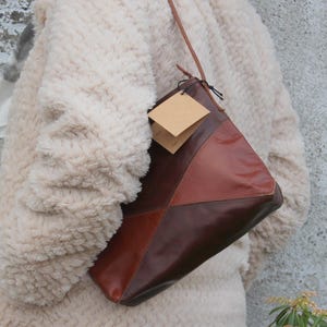 May include: A brown and tan leather shoulder bag with a geometric design. The bag has a long strap and a tag attached. The bag is being worn over a cream-coloured, textured coat.