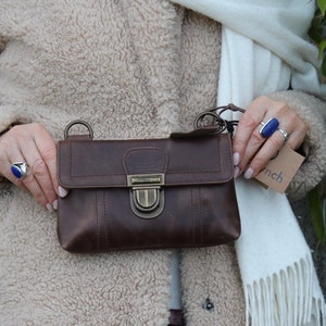Può includere: Una borsa a tracolla in pelle marrone con forma rettangolare e chiusura con fibbia in metallo. La borsa ha una tracolla e un piccolo cartellino attaccato. La borsa è tenuta davanti a un cappotto beige e una sciarpa bianca.