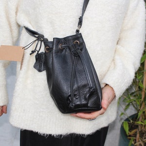 Puede incluir: Un bolso de cuero negro de estilo cubo con cierre de cordón y una correa larga. El bolso está siendo sostenido por una persona que lleva un suéter blanco.