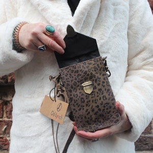May include: A small brown and black leopard print crossbody bag with a gold clasp and a black flap. The bag is being held by a person wearing a white faux fur jacket. The bag has a brown leather strap and a tag that says "Oddly Enough".