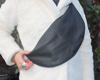 Puede incluir: Un bolso negro de cuero en forma de media luna que se lleva cruzado. El bolso lo sostiene una persona que lleva un abrigo blanco de piel sintética. El bolso tiene una superficie texturizada y un diseño curvo.