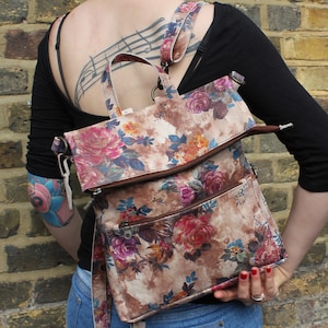 May include: A floral print leather backpack with a zippered flap and multiple pockets. The bag has a brown leather strap and is worn over the shoulder.