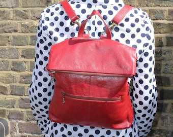 Puede incluir: Una mochila de cuero roja con cierre de solapa y un bolsillo con cremallera. La mochila la lleva puesta una persona que lleva una camisa y una falda de lunares blancos y negros.