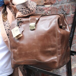 May include: A brown leather satchel with a clasp closure and a top handle. The bag has a vintage look with visible wear and tear. A tag with the text "Od Lynch" hangs from the handle. The bag is resting on a black metal structure against a brick wall.