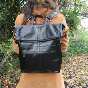 Puede incluir: Una mochila de cuero negro con una solapa abatible y un bolsillo con cremallera. La mochila la lleva puesta una persona con el pelo largo y castaño.