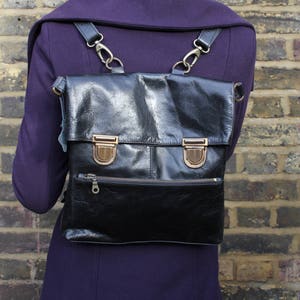 May include: Black leather backpack with brass-colored hardware, worn over a purple coat. The backpack has a flap closure with two buckles, a front zipper pocket, and adjustable straps. The bag is worn against a brick wall.
