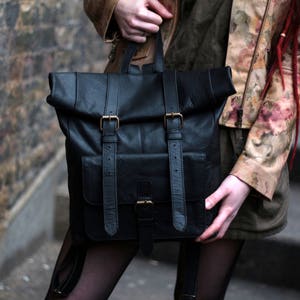 May include: Black leather backpack with a roll-top closure and two front pockets. The bag features adjustable straps and gold-colored buckles. The backpack is held by a person wearing a floral jacket.