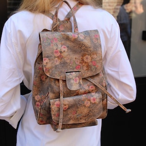 Puede incluir: Una mochila marrón con un estampado floral de rosas rosas y rojas. La mochila tiene un cierre de solapa, un bolsillo delantero y correas ajustables. La bolsa está hecha de un material texturizado.