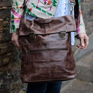 May include: A brown leather satchel bag with a flap closure and two metal buckles. The bag has a zipped pocket on the front and a strap for carrying.
