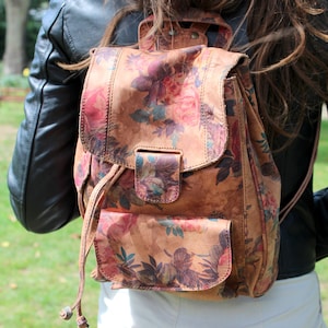 Puede incluir: Una mochila de cuero marrón con un estampado floral de rosas rosas y moradas y hojas verdes. La mochila tiene un cierre de solapa, un bolsillo delantero y correas ajustables. Se lleva sobre una chaqueta de cuero negra.