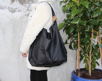 May include: A large, black leather tote bag with a shoulder strap. The bag is worn over the shoulder. The person is wearing a white coat. A plant in a blue pot is in the background.