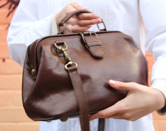 May include: A brown leather doctor's bag with a top handle and a shoulder strap. The bag has a brass buckle and clasp.