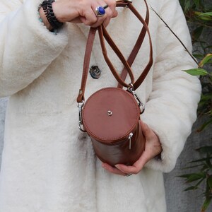 May include: A brown leather cylindrical crossbody bag with a silver zipper and hardware. The bag has a circular top and bottom and is held by a person wearing a white coat. The bag has a long, thin strap.