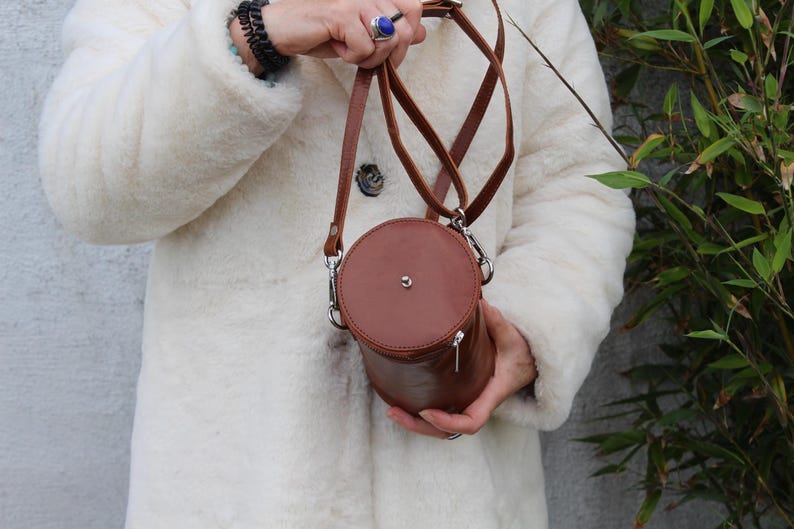 May include: A brown leather cylindrical crossbody bag with a silver zipper and hardware. The bag has a circular top and bottom and is held by a person wearing a white coat. The bag has a long, thin strap.