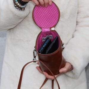 May include: A brown leather crossbody bag with a circular top and cylindrical body. The interior is lined with pink fabric featuring white polka dots. The bag is open, revealing a phone and a wallet. A silver strap and zipper are visible.