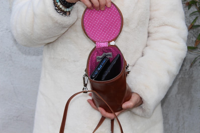 May include: A brown leather crossbody bag with a circular top and cylindrical body. The interior is lined with pink fabric featuring white polka dots. The bag is open, revealing a phone and a wallet. A silver strap and zipper are visible.