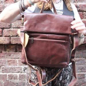 May include: A brown leather satchel style backpack with a flap closure and multiple pockets. The bag has a long adjustable strap.
