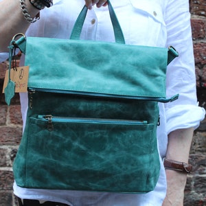 May include: A teal leather backpack with a flap closure and two zipped pockets. The bag has a brown leather strap and a small brown tag with the text "Del's Dye" on it.
