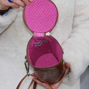 May include: A brown leather cylindrical handbag with a zipper closure. The interior is lined with pink fabric and white polka dots. A small tag is visible inside the bag. The bag has a shoulder strap.