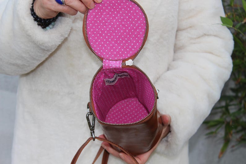 May include: A brown leather cylindrical handbag with a zipper closure. The interior is lined with pink fabric and white polka dots. A small tag is visible inside the bag. The bag has a shoulder strap.