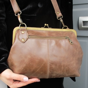 May include: A brown leather crossbody bag with a gold clasp and a zipped pocket. The bag has a long strap and is made of soft, supple leather.