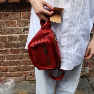 Puede incluir: Una ri&ntilde;onera de cuero roja con un bolsillo con cremallera y una correa ajustable. La bolsa la lleva una persona que lleva una camisa blanca y pantalones de color claro.