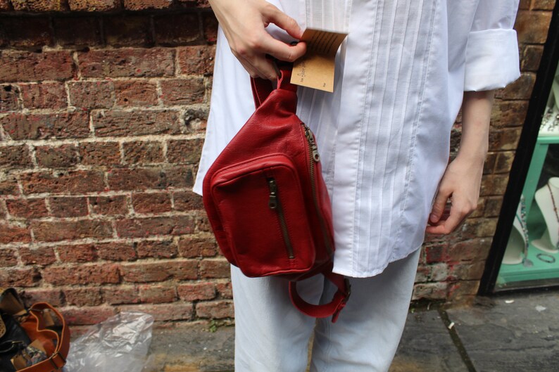 Puede incluir: Una ri&ntilde;onera de cuero roja con un bolsillo con cremallera y una correa ajustable. La bolsa la lleva una persona que lleva una camisa blanca y pantalones de color claro.