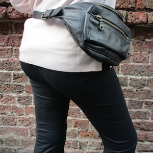 May include: A black leather fanny pack with a zipper closure and a silver buckle. The fanny pack is worn around the waist of a person wearing a pink sweater and black pants.