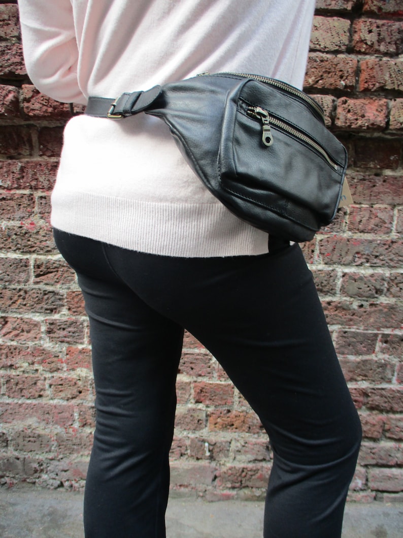 May include: A black leather fanny pack with a zipper closure and a silver buckle. The fanny pack is worn around the waist of a person wearing a pink sweater and black pants.