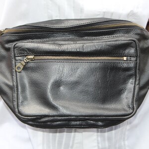 May include: Black leather fanny pack with multiple zippered compartments and a front pocket. The bag has gold-colored zippers and hardware. The leather appears to be slightly textured. The bag is held up by a person wearing a white shirt.