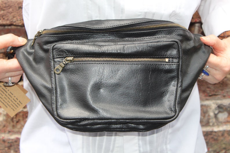 May include: Black leather fanny pack with multiple zippered compartments and a front pocket. The bag has gold-colored zippers and hardware. The leather appears to be slightly textured. The bag is held up by a person wearing a white shirt.