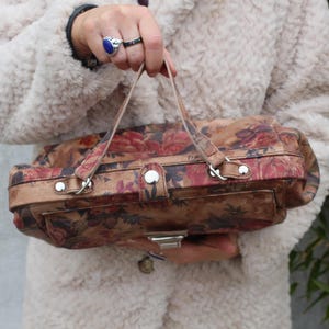 May include: A vintage handbag featuring a floral design in red, pink, and brown hues. The rectangular bag has a top handle and a silver clasp. The background is a cream-coloured coat.