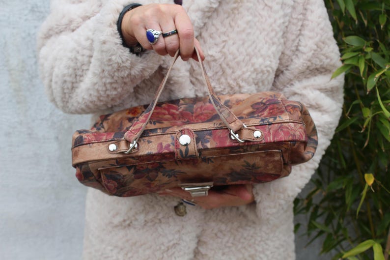 May include: A vintage handbag featuring a floral design in red, pink, and brown hues. The rectangular bag has a top handle and a silver clasp. The background is a cream-coloured coat.