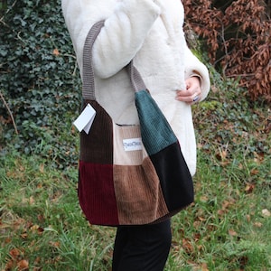 Puede incluir: Bolso de hombro de pana con diseño patchwork en tonos marrón, burdeos, verde azulado y negro. El bolso tiene forma rectangular con una correa larga y una pequeña etiqueta adjunta. Una persona con un abrigo blanco sostiene el bolso.