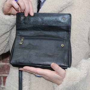 K&ouml;nnte beinhalten: Eine schwarze Leder-Umh&auml;ngetasche mit einer Rei&szlig;verschlusstasche und einem Klappverschluss. Die Tasche ist offen und das Innere ist sichtbar.